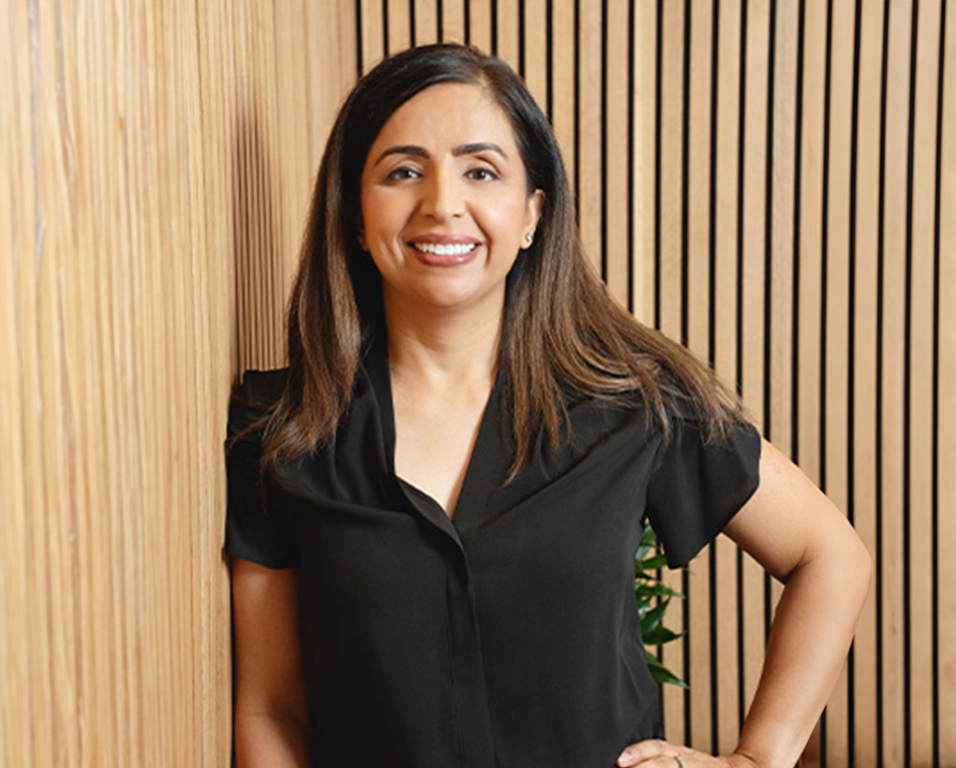 The image shows a woman standing against a wooden wall with vertical slats, smiling at the camera, wearing a black blouse, and has long dark hair.