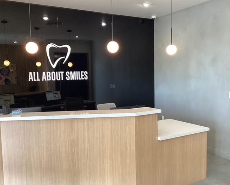 The image shows an interior view of a dental office reception area with a modern design, featuring a large window sign for  All About Smiles,  a company logo, and a white desk with a lamp.