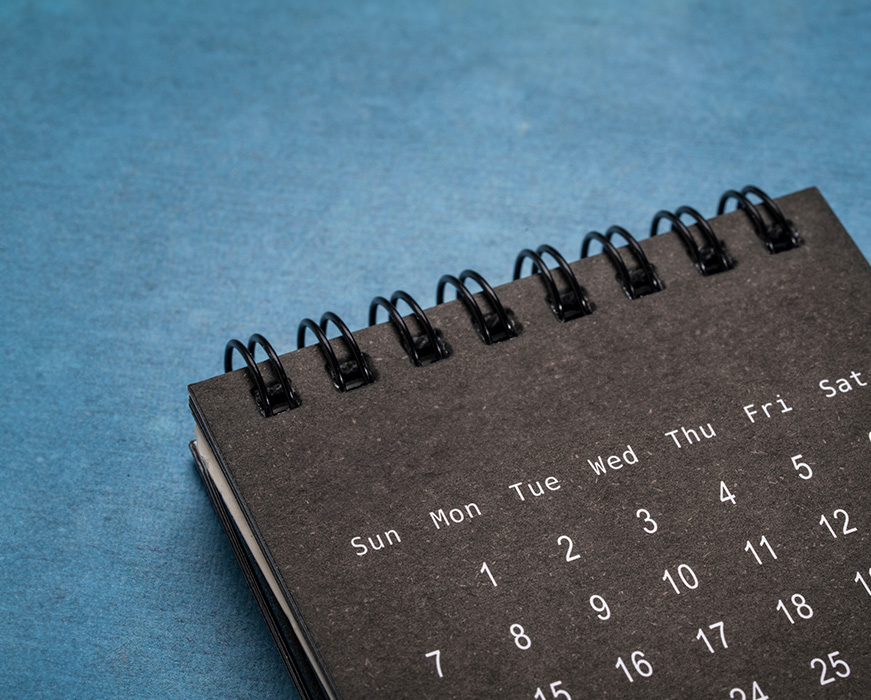 The image shows a black spiral-bound notebook with a calendar page open, displaying dates and times, placed on a blue surface.