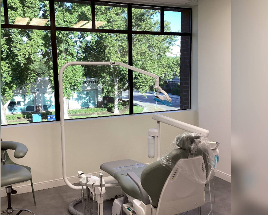 The image shows an interior view of a dental office with a large dental chair in the center, under a window with sunlight streaming in, and a dental mirror attached above the chair.