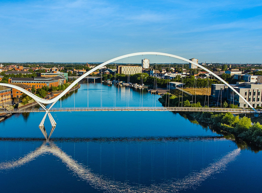 The image features a modern white arch bridge spanning across a calm body of water with a city skyline visible in the background, under a clear blue sky.