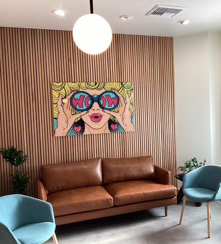 This image features a modern interior space with a large, colorful artwork depicting a stylized female figure wearing sunglasses and the word  Wow  on her forehead, mounted on a wall with a horizontal wood panel design. The room has a contemporary aesthetic with clean lines and minimal decorations, including a sofa, chairs, and a round pendant light.